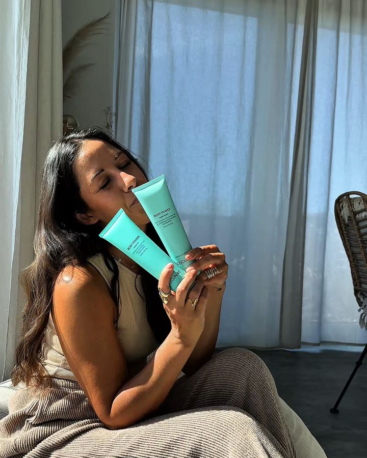 Creatorin hält zwei türkise Body-Gel-Tuben ans Gesicht, sonniges Wohnzimmer